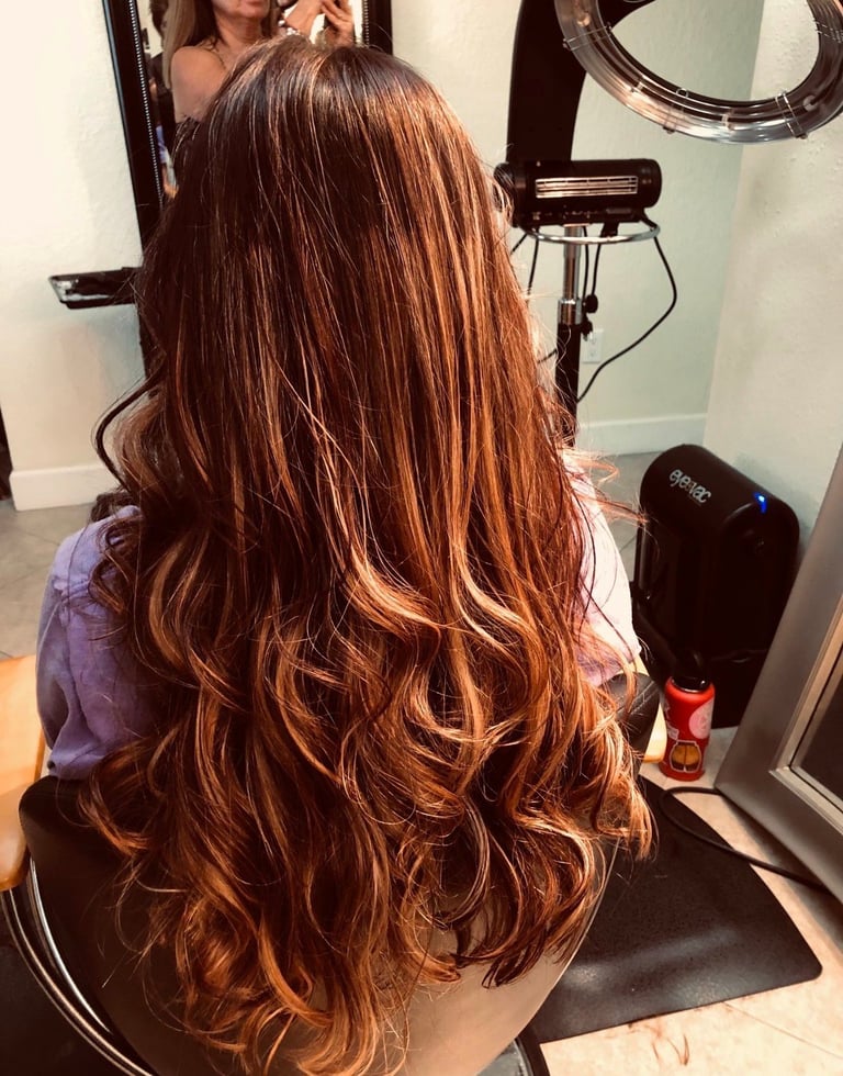 Woman with long copper-brown hair with highlights, shown from behind in a salon setting with mirrors and styling products
